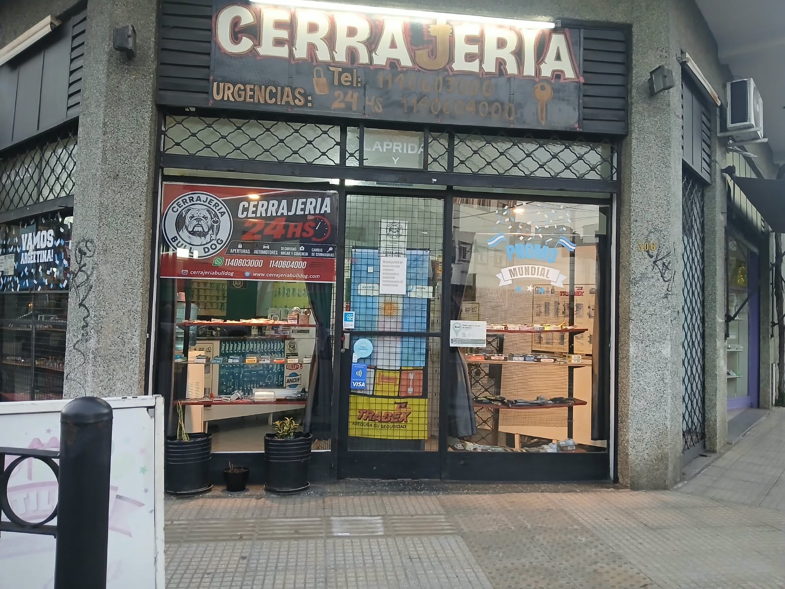 Fachada nocturna de Cerrajería Bulldog 24HS en Villa Martelli — cartel iluminado CERRAJERÍA, vidriera con productos